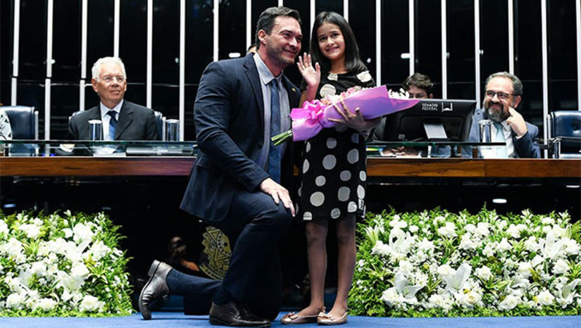 Imagem 1 -  Durante a sessão, a vida de Tiffany Brito, de 8 anos, foi celebrada. Ela recebeu flores das mãos do senador Styvenson Valentim (Podemos-RN)  e retribuiu o presente com um desenho em que a menina aparece ao lado do senador.  "Quero te homenagear em nome de todas as crianças que estão lutando contra o câncer", disse o senador Styvenson Valentin, que propôs e presidiu a sessão no Senado Federal em homenagem as vítimas do câncer. Na ocasião, defendeu o tratamento precoce.